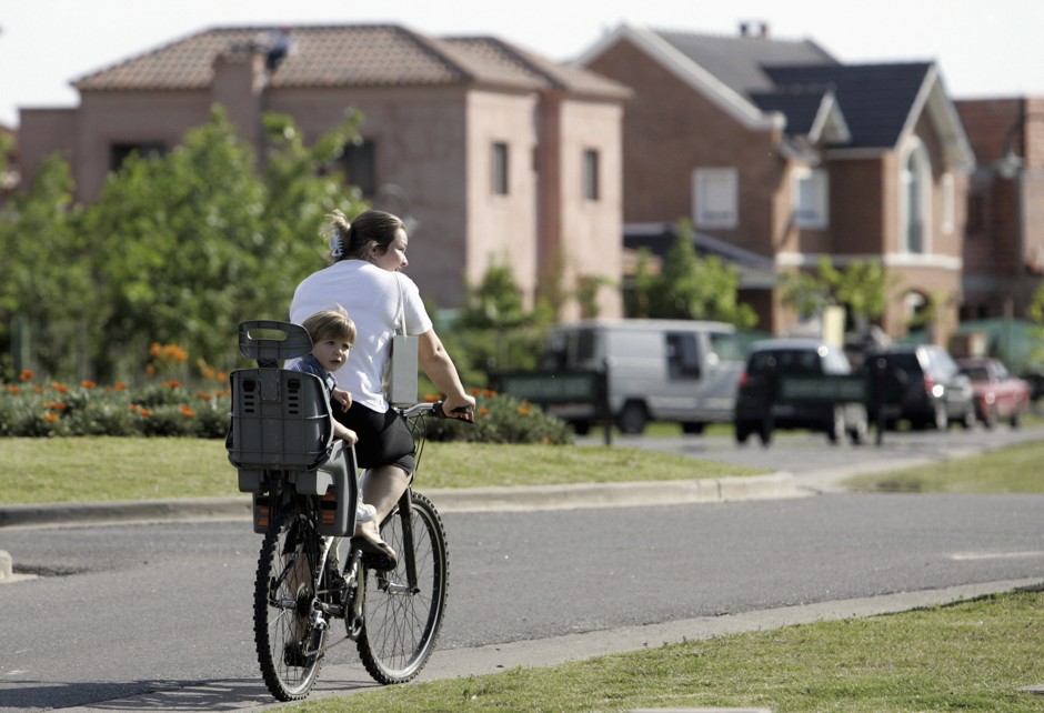 Two year-old Manuel and his baby-sitter Suni ride a bicycle in their neighbourhood at Nordelta gated community in Greater Buenos Aires, November 29, 2006. Since 1994, an estimated 300.000 people have fled high-density, high-crime, high-priced Buenos Aires and its traditional suburbs near the city core, to live far on the outskirts in pastel houses along idyllic artificial lakes, with 24-hour security guards screening visitors at the perimeter fence. Picture taken on November 29, 2006. To match feature CITIES/ARGENTINA-GATED REUTERS/Marcos Brindicci (ARGENTINA) - RTR1KAPD